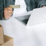 Person using a credit card for online shopping on a laptop surrounded by shipping boxes.