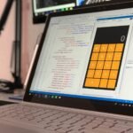 A laptop screen shows a coding application with a calculator design in a tech office setting.