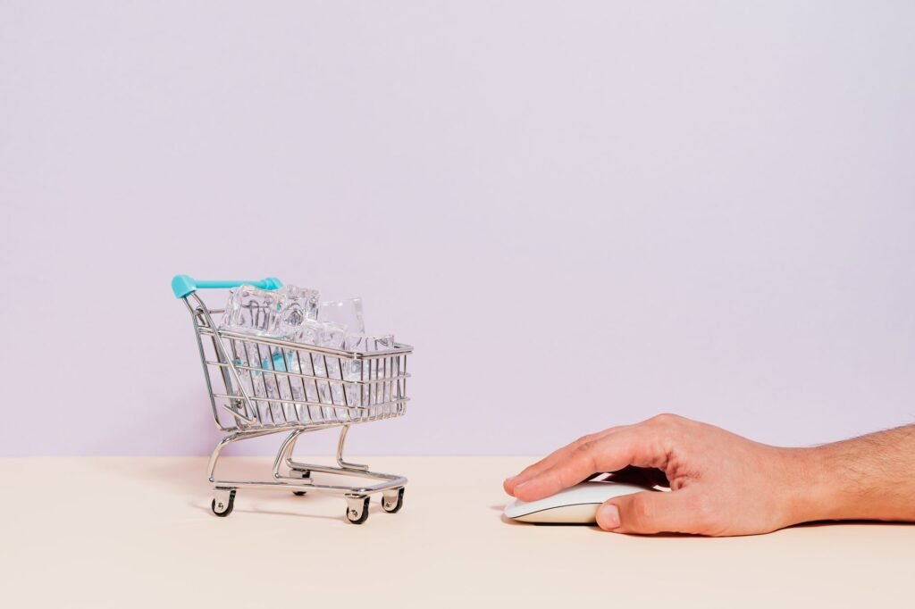Conceptual image of a hand on a mouse next to a miniature shopping cart filled with ice cubes, symbolizing online shopping.