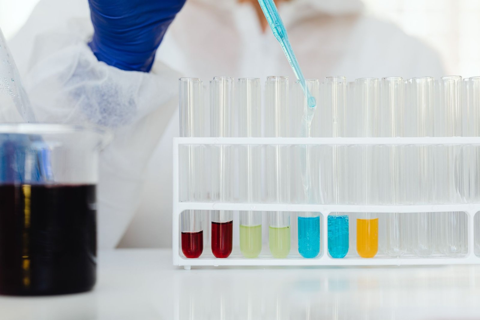Scientist in protective gear using a dropper to fill test tubes with colorful liquids in a laboratory.