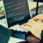 Close-up view of a developer typing code on a keyboard with a computer screen showing scripts.