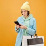 Trendy young woman using smartphone and holding shopping bags against yellow background.