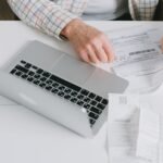 Close-up of hands organizing documents on a desk with a laptop.
