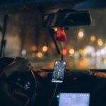 Driving at night in a car with GPS navigation on smartphone, city lights reflecting on wet windshield.