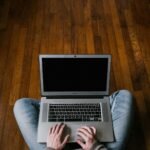 Person sitting cross-legged on hardwood floor using a laptop, focusing on minimalism.