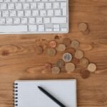 Scattered coins on a wooden desk alongside a keyboard and notebook, captured from above.