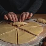 Close-up of hands rolling dough into croissants on a floured surface.